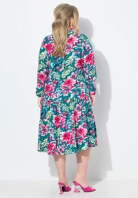 Vestido floral con base en tono verde azulado, grandes flores rosas y verdes, mangas largas y falda de volantes. Combinado con zapatos de tacón rosas.