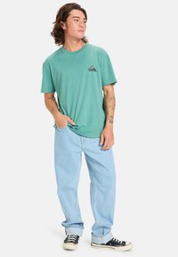 Turquoise cotton t-shirt with a black logo on the left chest, paired with light blue baggy jeans and black sneakers with white soles.