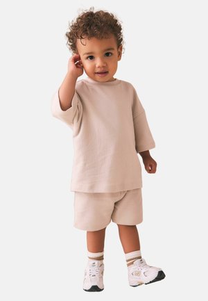 Väikelaps lokkis juustega, kannab beežikat oversize särk ja lühikesi pükse, valgeid tossusid ning triibulisi sokke, seisab käega kõrv lähedal.