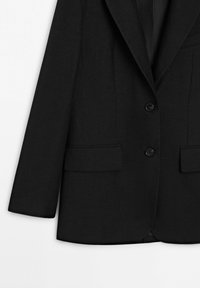 Zwarte formele blazer met twee knopen, twee klepzakken aan de voorkant en lange mouwen tegen een lichte achtergrond.