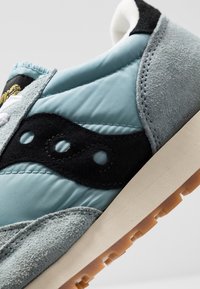 Gros plan sur une chaussure de sport en daim bleu clair et gris avec un logo noir superposé et une semelle beige à bande de roulement marron texturée.