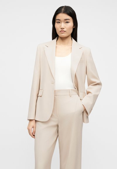 Γυναίκα που φορεί κοστούμι σε μπεζ χρώμα, με σακάκι και παντελόνι που ταιριάζουν, συνδυασμένο με λευκό ριμπ αμάνικο τοπ, να στέκεται μπροστά σε απλό φόντο.