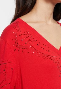 Suéter rojo con cuello en V, adornado con strass decorativos en un patrón en los hombros. Textura suave con bordes acanalados.