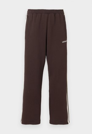 Uomo che indossa pantaloni della tuta ampi color marrone scuro con strisce laterali e sneakers bianche, in piedi davanti a uno sfondo bianco.