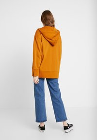 Sudadera con capucha naranja de corte relajado, que llega por debajo de las caderas, combinada con unos vaqueros azules holgados y unos zapatos negros sin cordones.