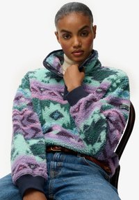 Pull polaire en vert menthe, lavande et marine avec motifs géométriques, col haut et poignets côtelés ; logo sur la manche ; porté avec un jean en denim.