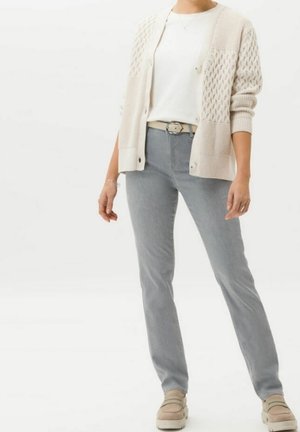 Cardigan en maille beige clair avec motif texturé, porté sur une chemise blanche, associé à un jean gris clair et des chaussures beige à enfiler.