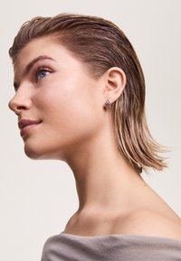 Orecchini con un design a scacchi in argento, con piccoli accenti di pietre preziose, indossati da una persona con capelli corti e pettinati all'indietro.