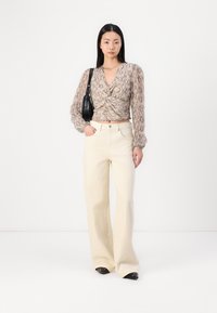Blusa a maniche lunghe con motivo a serpente in marrone chiaro, annodata in vita, abbinata a pantaloni larghi crema e calzature nere, con una borsa nera.