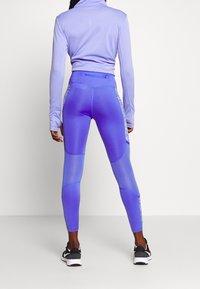 Fialový sportovní outfit obsahující top s dlouhým rukávem a těsné legíny. Legíny mají gradientní design a síťové panely. Viditelné černé tenisky.