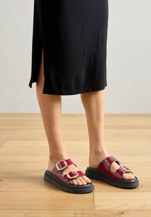 Rode en zwarte slipper sandalen met twee gesps, textuur bovenkant en een zwarte rubberen zool. G gedragen met een zwarte jurk en houten vloer als achtergrond.