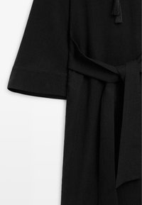 Vêtement en tissu noir avec une manche large, ceinture nouée à la taille, et texture du tissu visible sur un fond blanc.