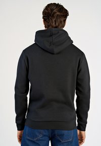 Svart hoodie tillverkad av mjukt material, har en avslappnad passform, ribbade muddarna och nederkanten, samt en uttalad huva. Bärs med blå jeans.