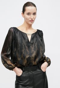 Schwarze transparente Bluse mit einem goldenen Schlangenmuster, einem Ausschnitt mit dekorativer Metallverzierung und weiten Ärmeln. Kombiniert mit schwarzen Lederhosen.