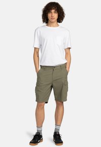 Pantalones cortos cargo en color verde oliva con múltiples bolsillos, combinados con una camiseta blanca lisa. El modelo lleva zapatillas negras y calcetines a rayas.