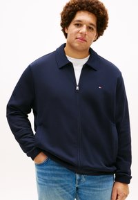 Μπλε ναυτικό μπουφάν με φερμουάρ, γιακά, απαλής υφής και με έμβλημα λογότυπου στο αριστερό στήθος. Συνδυάζεται με ανοιχτόχρωμο τζιν παντελόνι.