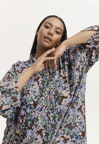 Kwiatowa koszula w brązowym kolorze z niebieskimi i zielonym akcentami; ma wysoki kołnierz, zapięcie na guziki z przodu oraz luźne rękawy.