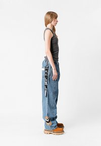 Vaqueros de denim azules holgados con un parche de oso, una camiseta sin mangas gris y botas color beige. Incluye un bolsillo cargo visible y detalles de correa con marca.