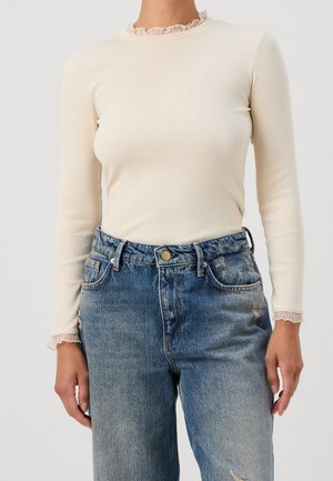 Vrouw draagt een beige geribd topje met lange mouwen en gerimpelde kraag en manchetten, gecombineerd met een hooggesloten vervaagde blauwe jeans.