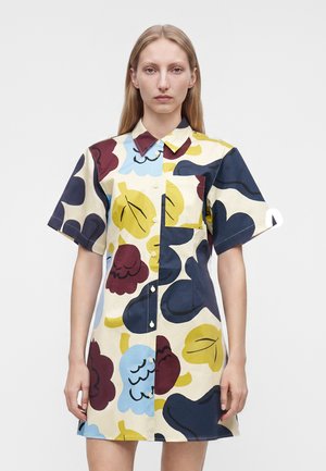 Vrouw draagt een kortemouw button-up jurk met meerkleurige abstracte bloemmotieven in geel, blauw, bordeauxrood en marineblauw.