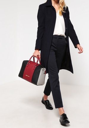 Nainen kävelee mustassa takissa, valkoisessa paidassa, mustissa housuissa ja mustissa kengissä, kantaen suurta punaista, mustaa ja harmaata käsilaukkua.