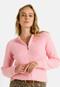 Cardigan zippé rose en tricot avec une texture duveteuse, des poignets côtelés et un col arrondi. Doté d'une fermeture éclair argentée. Porté avec un bas à motifs.