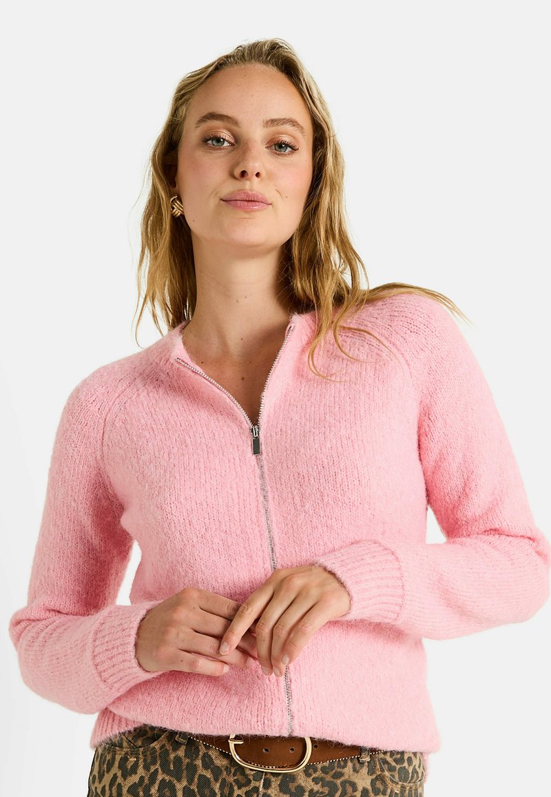 Cardigan zippé rose en tricot avec une texture duveteuse, des poignets côtelés et un col arrondi. Doté d'une fermeture éclair argentée. Porté avec un bas à motifs.