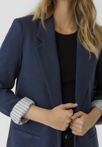 Granatowa marynarka z klapami z wcięciem, z paskiem na mankiecie podwiniętych rękawów i ukrytym zapięciem z przodu. Gładka faktura tkaniny.