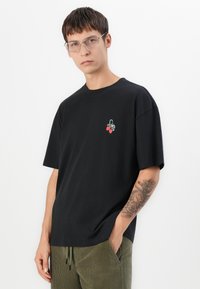 Sort bomulds T-shirt med et lille broderet design af jordbær. Korte ærmer, afslappet pasform og rund hals. Modellen har olivengrønne bukser på.