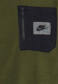 Olivově zelená fleecová látka s černou kapsou na zip, která obsahuje skvrnitou záplatu s logem Nike pro detail a branding.