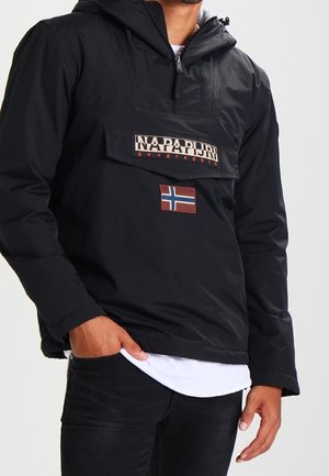 Mann trägt schwarze Napapijri-Jacke mit halbem Reißverschluss, Fronttasche und norwegischem Flaggenaufnäher, kombiniert mit schwarzen Hosen und weißem Hemd darunter.
