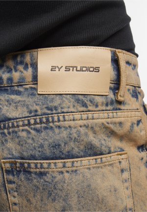 Bagsiden af blå og beige syrevaskede jeans med en tan læderlabel, der læser "2Y STUDIOS" over baglommen, båret med en sort top.