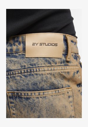 Bagsiden af blå og beige syrevaskede jeans med en tan læderlabel, der læser "2Y STUDIOS" over baglommen, båret med en sort top.