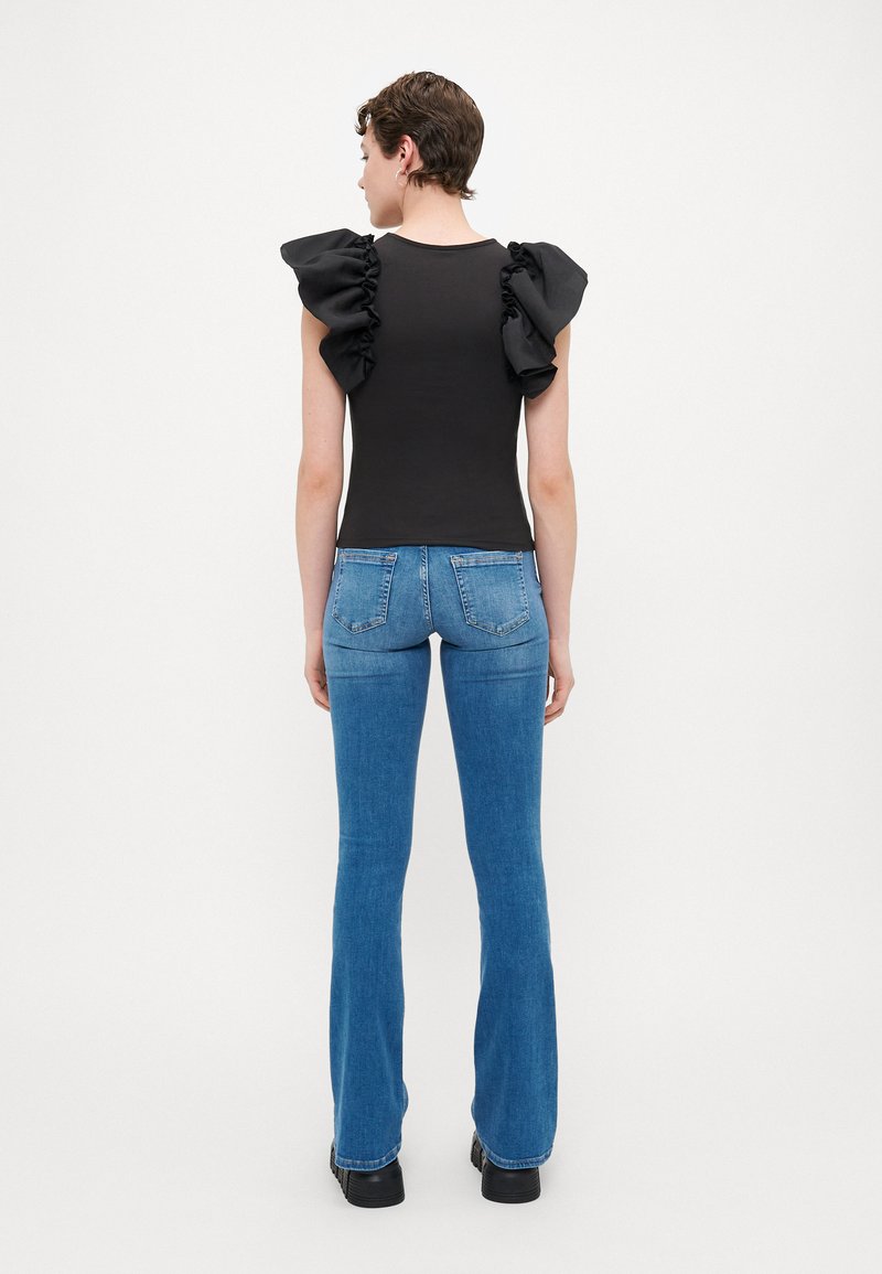 Schwarzes Top mit Rüschensleeves und blauen Schlagjeans. Das Model ist mit dem Rücken zur Kamera gewandt und zeigt das Design des Tops sowie die figurbetonte Silhouette der Jeans.