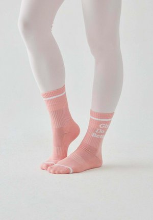 Nogi w białych rajstopach, noszące różowe skarpetki z białymi paskami i napisem „Girls Do Better” na jednej kostce na jasnym, jednolitym tle.