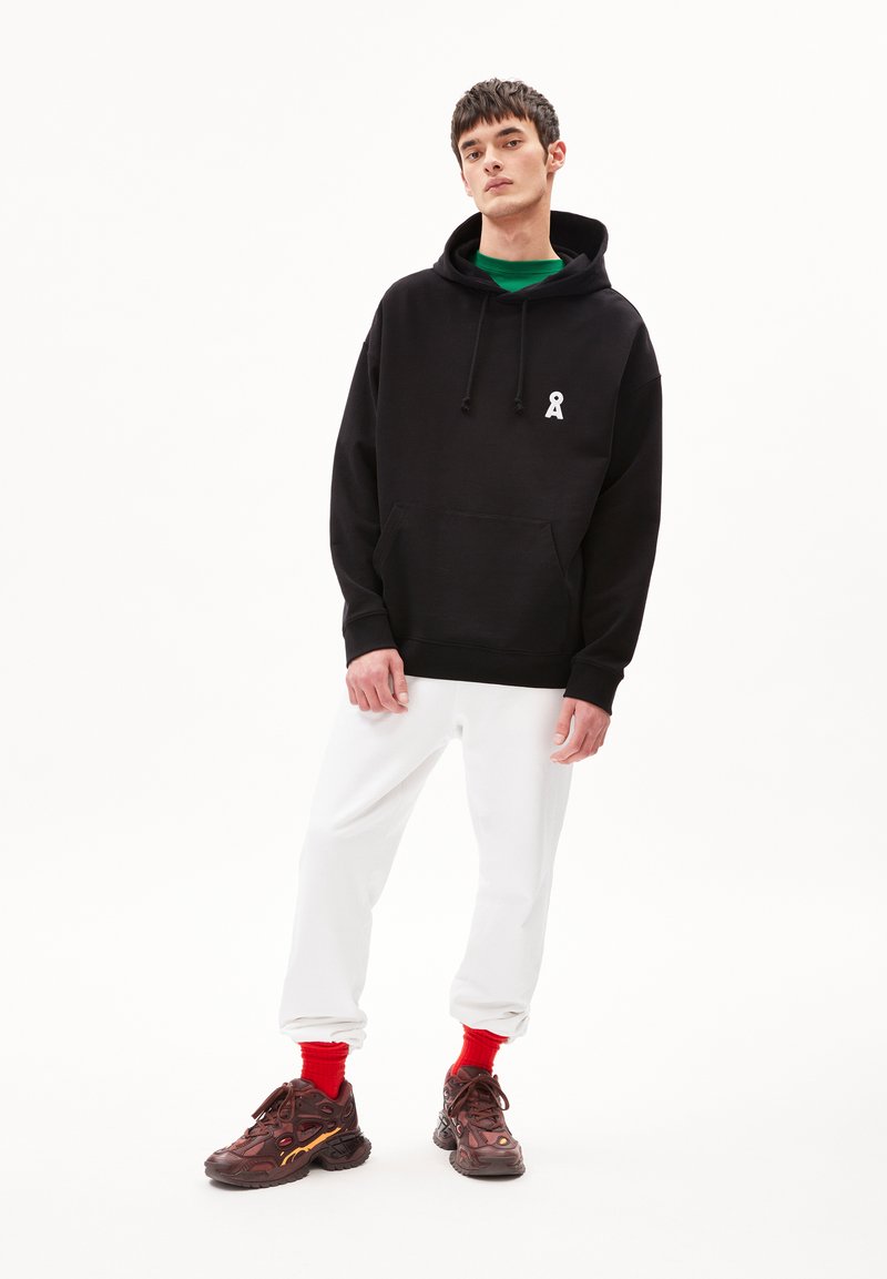 Felpa nera con cappuccio e logo bianco, abbinata a pantaloni bianchi larghi, calzini rossi e sneaker marroni con dettagli testurizzati.