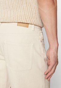 Vaqueros de mezclilla beige con una textura suave, que cuentan con un bolsillo trasero y una etiqueta de cuero en la cinturilla, mostrando un diseño minimalista.