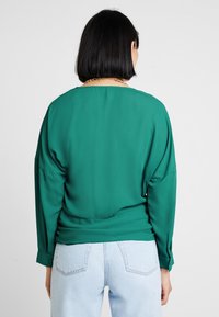 Person mit glattem schwarzen Haar, der eine grüne Langarmbluse und hellblaue Jeans trägt, mit dem Rücken zu einem schlichten Hintergrund.