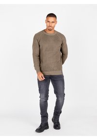 Olivgrüner gerippter Strickpullover mit Rundhalsausschnitt, kombiniert mit dunkler Jeans und schwarzen Schnürstiefeln. Schlichter Hintergrund, stehende Pose.