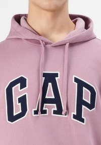 Pink hoodie with a drawstring and textured interior. "GAP" logo prominently displayed in navy with white outlining.