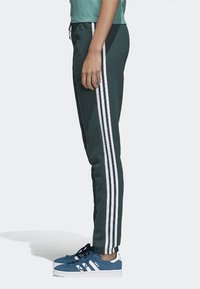 Dark green athletic pants with three white stripes down the sides, featuring a tapered fit and elastic waistband. Worn with blue sneakers.