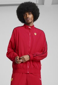 Rdeča atletska zadrgo jakna z visokim ovratnikom, črnima stranskima črtama in logotipom Ferrari na prsih. Lahka, gladka tekstura, elastični manšeti.