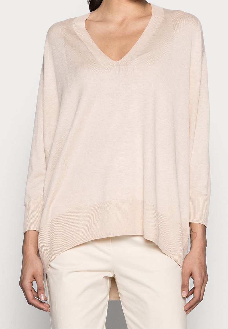 Kvinna iklädd en lös ljusbeige V-ringad tröja med långa ärmar och böjd nederkant, kombinerad med off-white byxor.