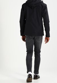 Czarna wodoodporna kurtka z kapturem, noszona na szarych obcisłych dżinsach. Posiada solidny pasek z tyłu oraz subtelne detale marki przy krawędzi.