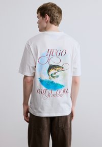Λευκό βαμβακερό T-shirt που απεικονίζει ένα μεγάλο γραφικό με ένα ψάρι που σπεύδει από μπλε κύματα, με κόκκινο κείμενο σε καλλιγραφική γραφή που γράφει "HUGO Bait 'N' Tackle Outfitters" στην πλάτη.
