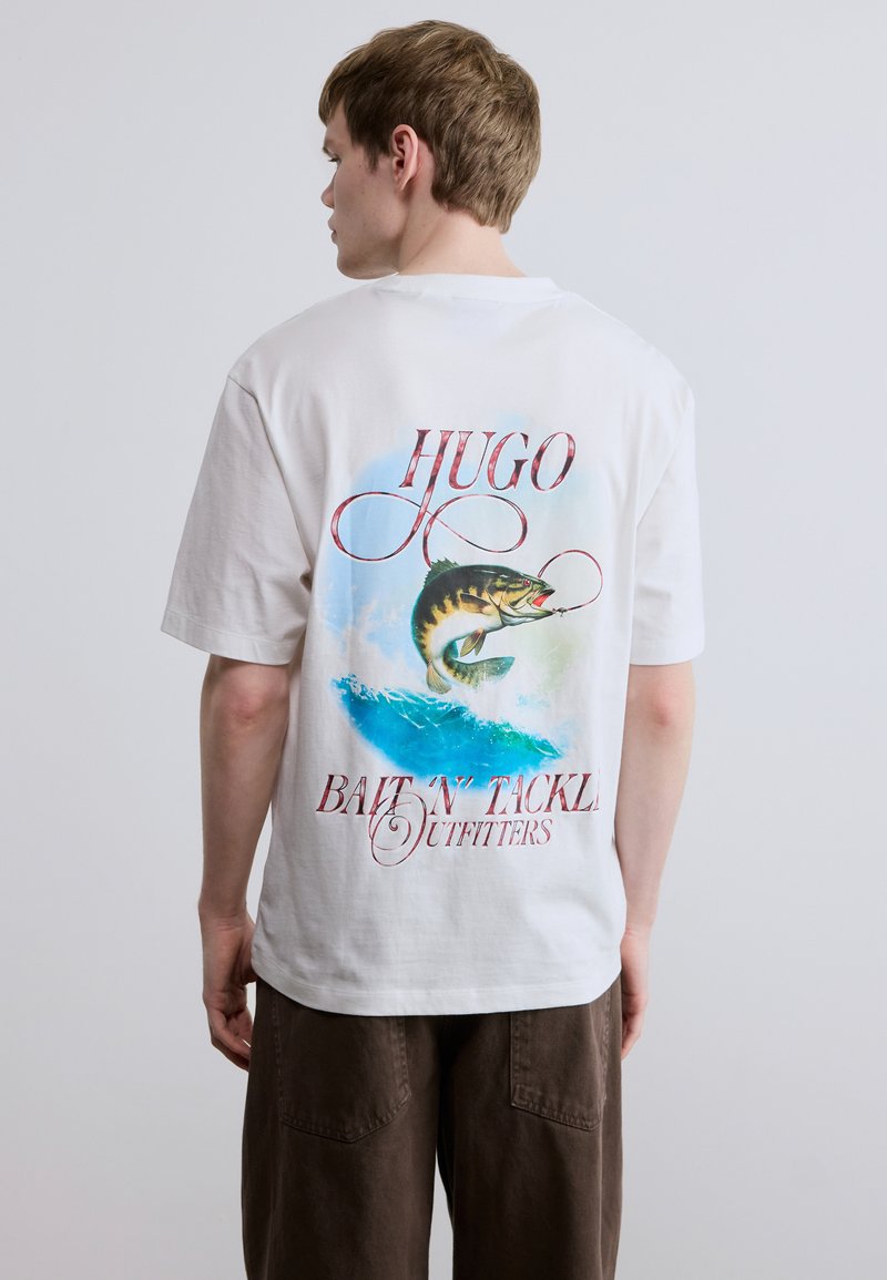 Λευκό βαμβακερό T-shirt που απεικονίζει ένα μεγάλο γραφικό με ένα ψάρι που σπεύδει από μπλε κύματα, με κόκκινο κείμενο σε καλλιγραφική γραφή που γράφει "HUGO Bait 'N' Tackle Outfitters" στην πλάτη.
