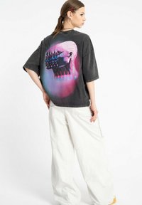 Przesadnie duża czarna koszulka z kolorową grafiką czaszki na plecach. Modelka połączyła ją z luźnymi białymi spodniami.