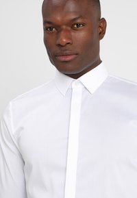 Hvit skjorte med strukturert krage, glatt tekstur og smal passform, med knapper foran og uten synlige mønstre.