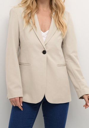 Femme portant un blazer beige à un bouton sur un top blanc avec un jean bleu, debout devant un fond clair uni.