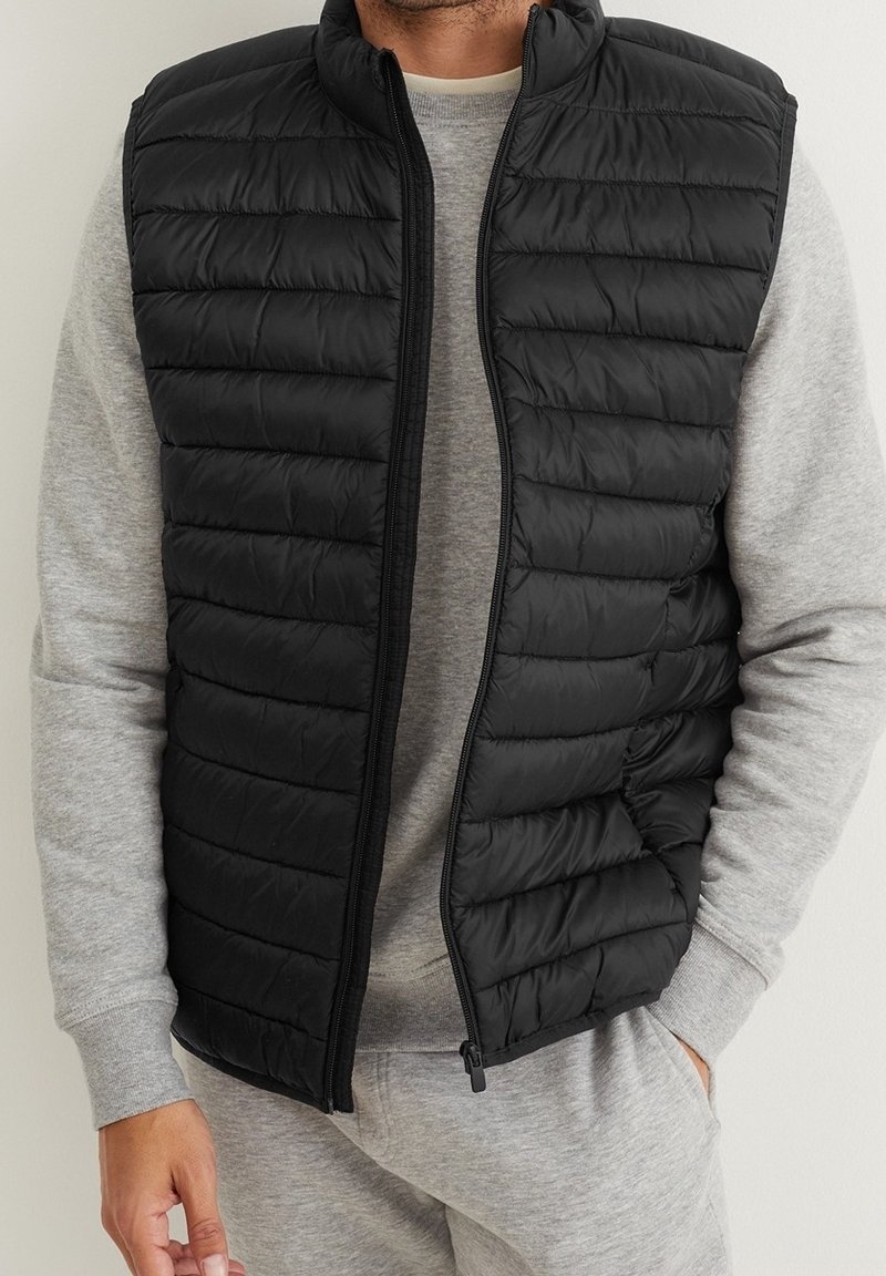 Czarna ocieplana kamizelka z zamkiem błyskawicznym, z poziomym przeszyciem i wysokim kołnierzem. Nosi się ją na szarą bluzę z prążkowanymi ściągaczami.